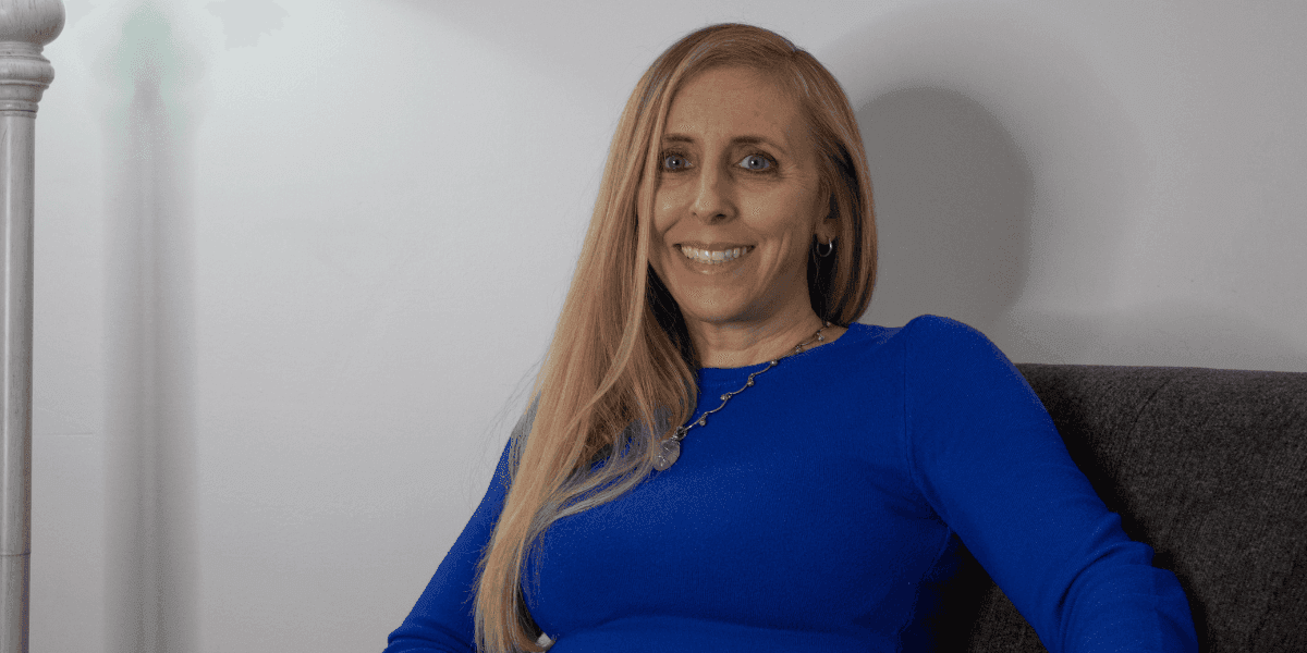 "Portrait of a smiling woman with long blonde hair, wearing a blue long-sleeve shirt, sitting comfortably on a gray couch, with soft lighting illuminating her face in a cozy indoor setting."