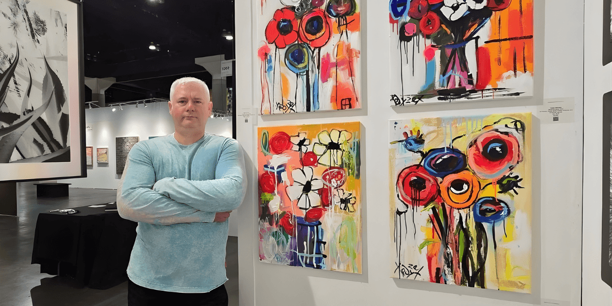 Man standing with arms crossed beside vibrant abstract flower paintings in contemporary art gallery.