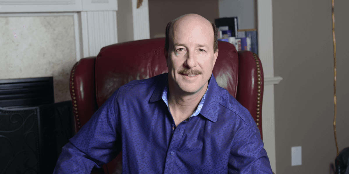 David Bissonette seated in red leather chair wearing purple shirt in home office setting.