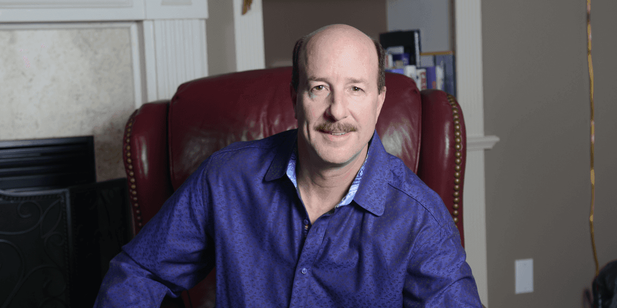 David Bissonette seated in red leather chair wearing purple shirt in home office setting.