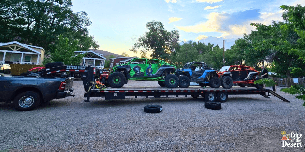 Off-road vehicles loaded onto a trailer for transportation in a scenic outdoor location with mountain views, featuring a green and blue dune buggy and an orange and black off-road vehicle.