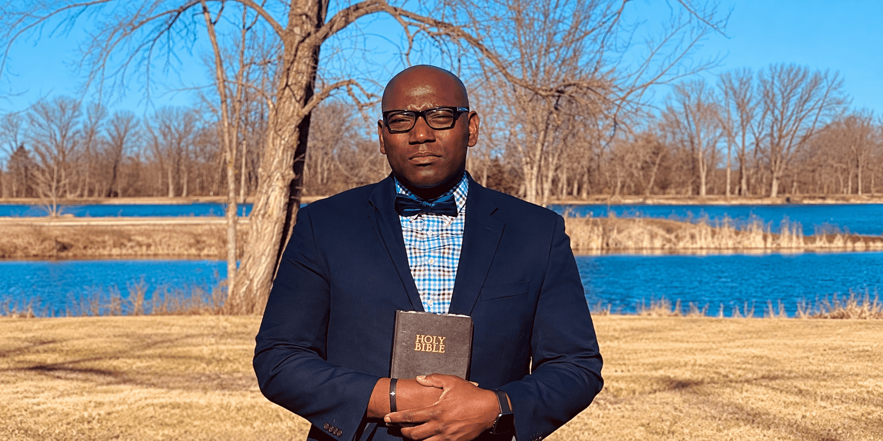 Brad D. Nelson standing outdoors by a lake holding a Holy Bible.