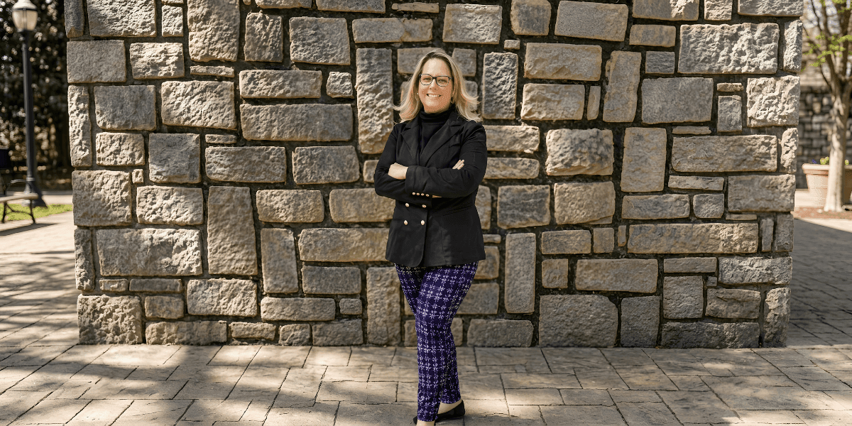 Professional woman standing with arms crossed in front of a stone wall outdoors, smiling confidently.