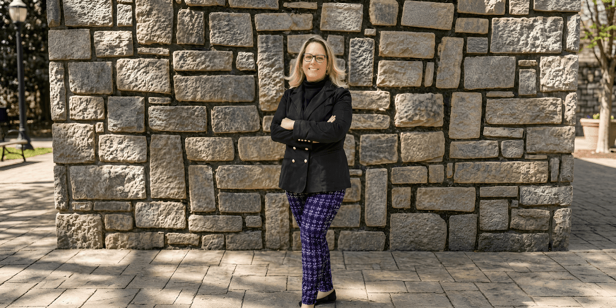 Professional woman standing with arms crossed in front of a stone wall outdoors, smiling confidently.
