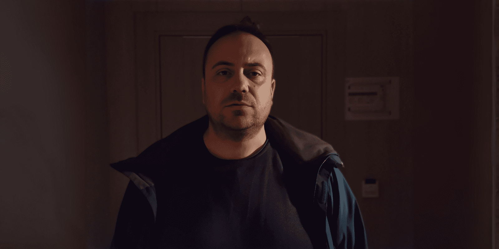 A man stands in a dim hallway, staring solemnly at the camera, wearing a dark t-shirt and jacket.