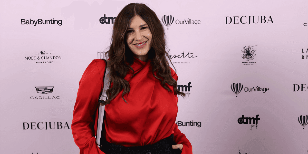 "Woman in a red blouse posing on the red carpet at a glamorous event, smiling at the camera with branded event logos in the background, showcasing her stylish look."