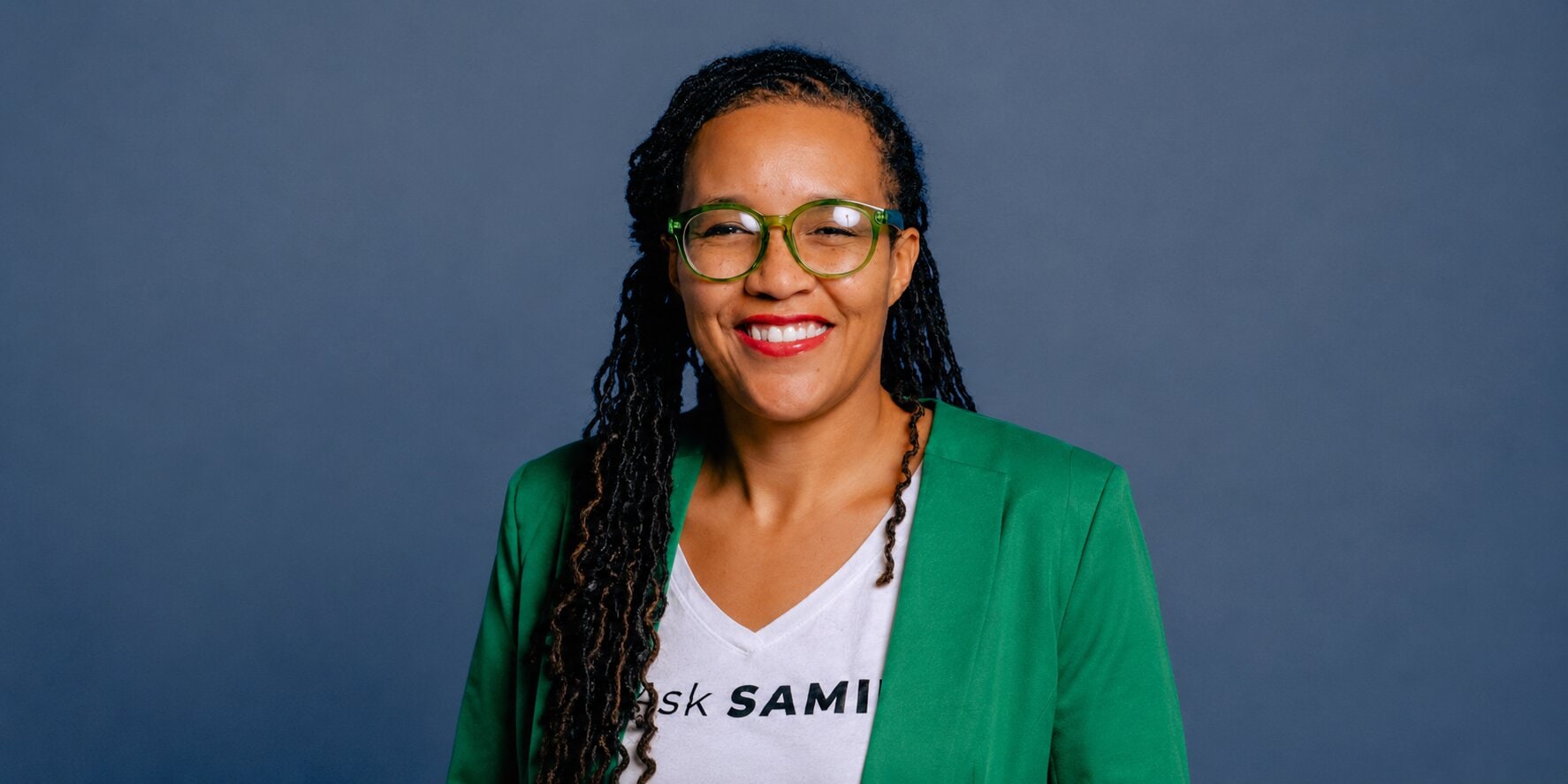 Dr. Brandy Archie smiling in green blazer and AskSAMIE shirt against a blue background.
