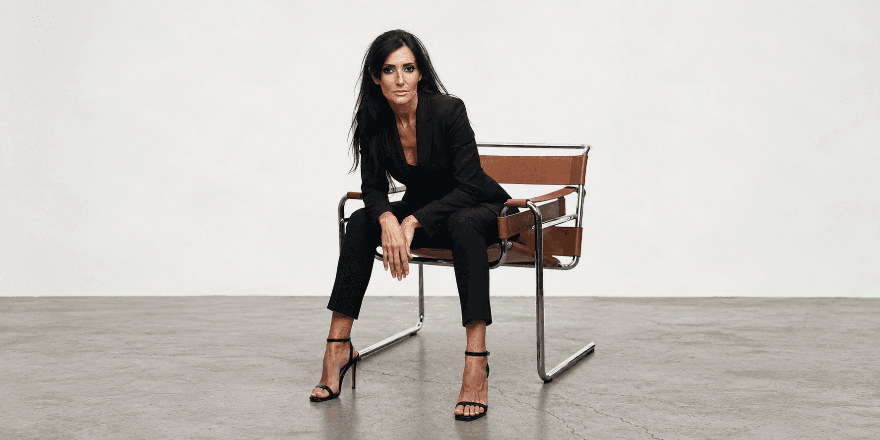 Silvia Rizzo seated in a modern chair wearing a black suit and high heels.