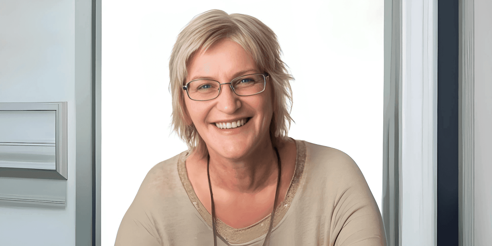 Smiling woman with short blonde hair, wearing glasses and a beige top, standing in front of a white door frame with a bright background.