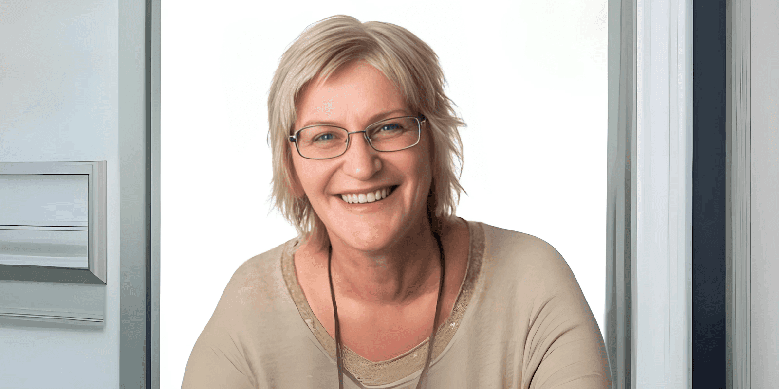 Smiling woman with short blonde hair, wearing glasses and a beige top, standing in front of a white door frame with a bright background.