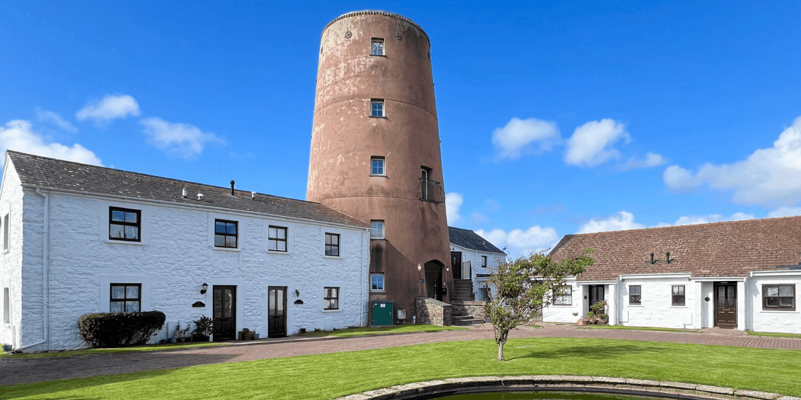 "A picturesque mill tower surrounded by white buildings and lush green grass on a clear day with blue skies and scattered clouds."