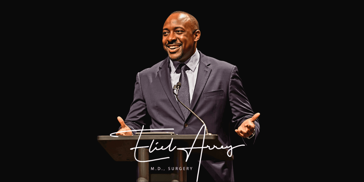 Dr. Eliel Arrey speaking at a podium in a suit against a black background.