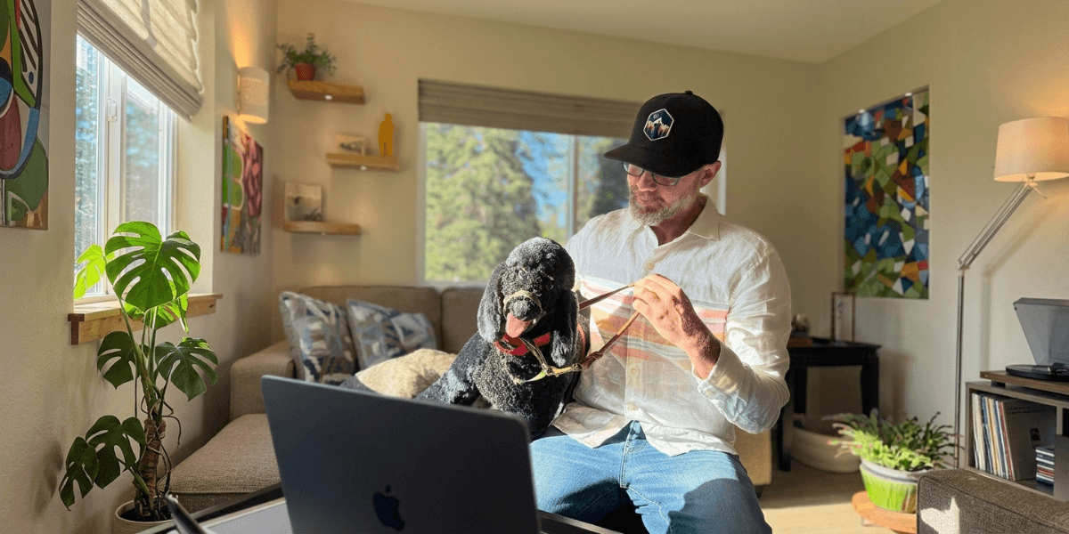 Blair Diamond sitting with a black dog beside a laptop in a sunlit living room.