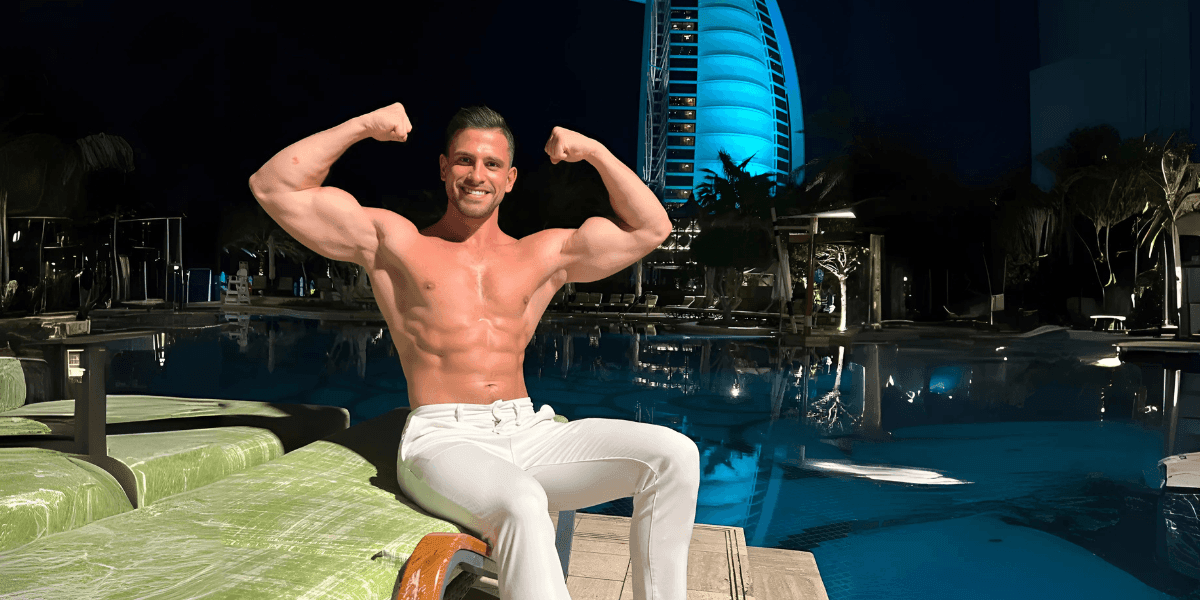 Shirtless man flexing by a pool at night with Burj Al Arab lit up in the background.