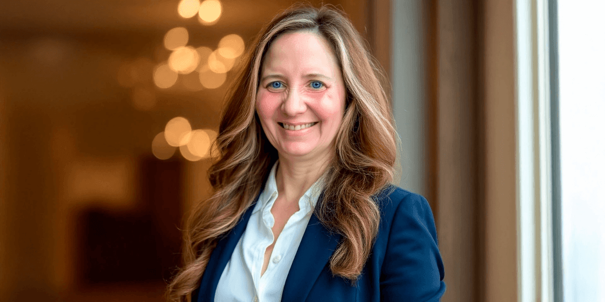 Sheera Eby, AI transformation consultant, smiling confidently in a professional blue jacket, with blurred lights in the background