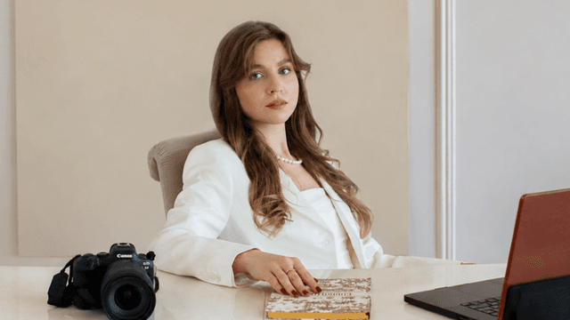 A woman in a white suit seated at a desk with a camera and laptop