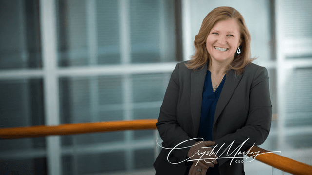 A smiling woman in a professional outfit leans against a railing indoors