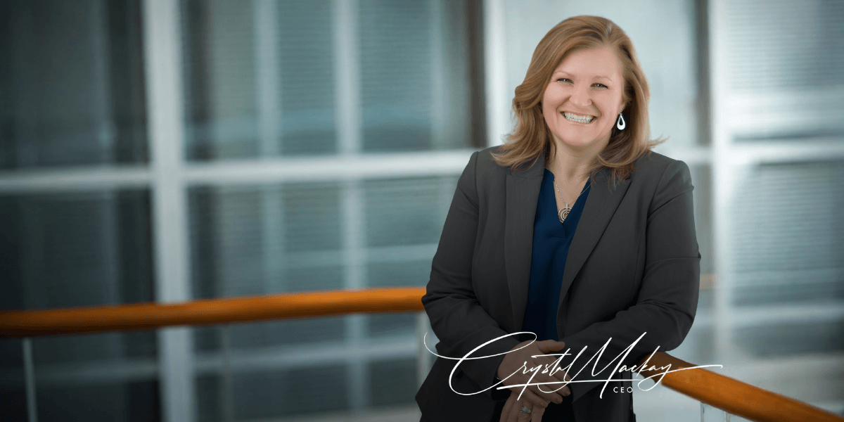 A smiling woman in a professional outfit leans against a railing indoors
