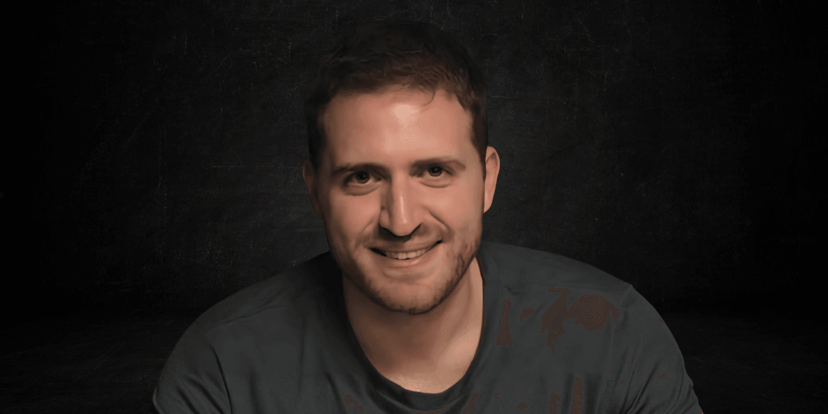 A smiling man with short brown hair, wearing a dark t-shirt, against a dark background
