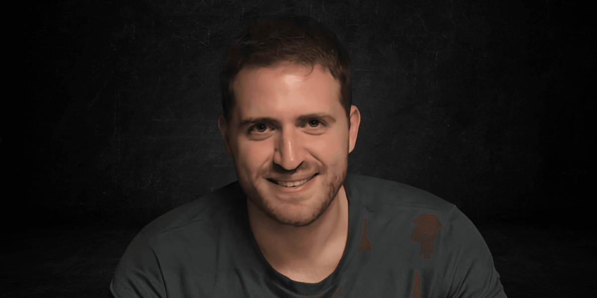 A smiling man with short brown hair, wearing a dark t-shirt, against a dark background