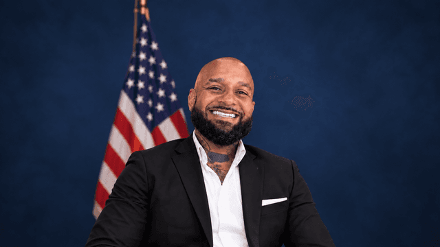 Smiling man in a black suit seated against an American flag background