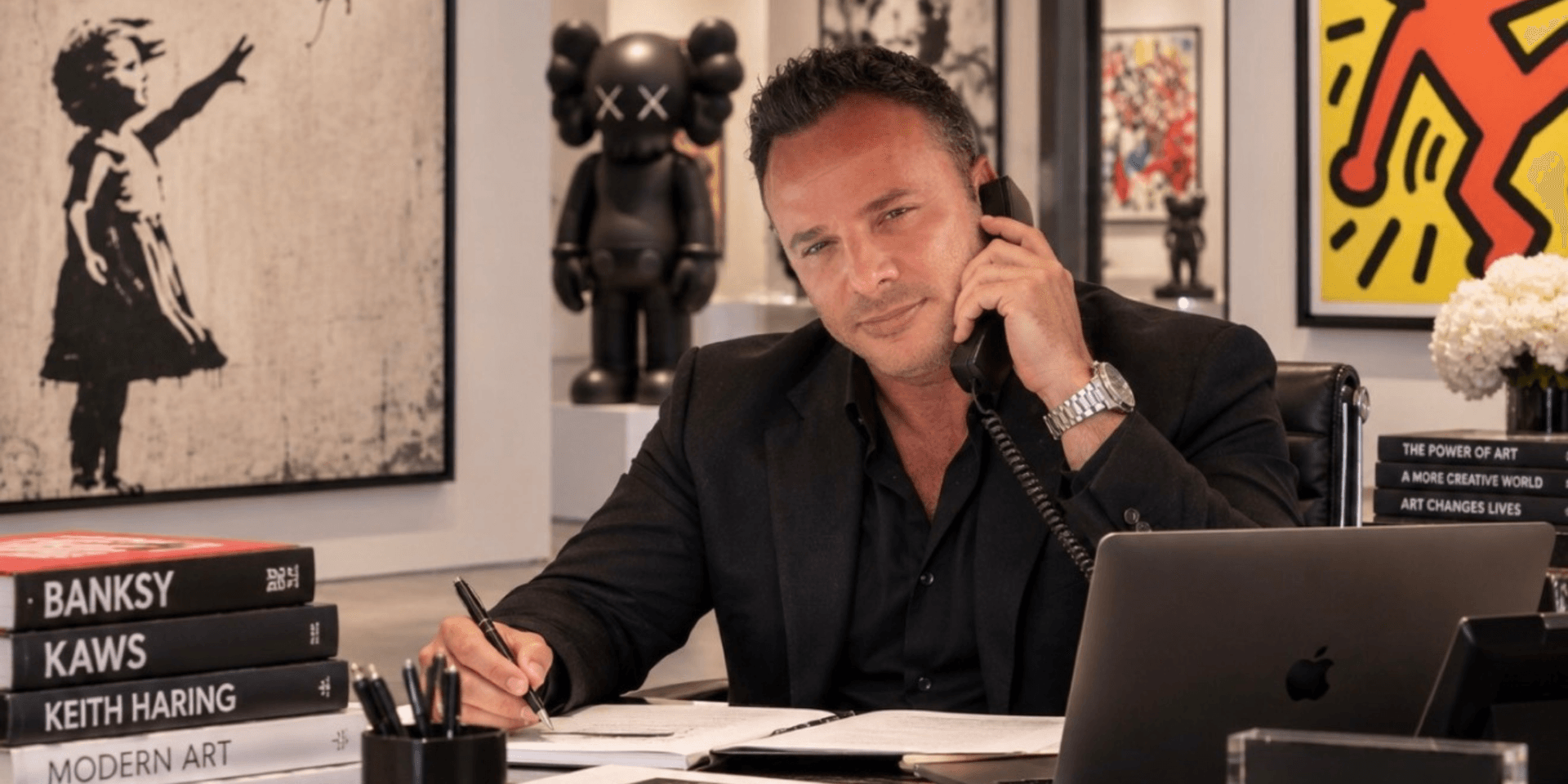 A man in a black suit sits at a desk, talking on the phone and writing.