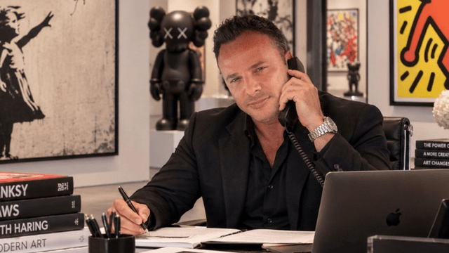 A man in a black suit sits at a desk, talking on the phone and writing.