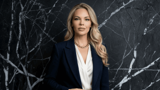 A woman in a blazer stands against a textured dark background with white patterns