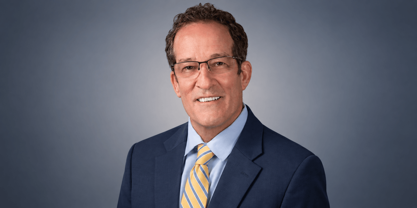 A middle-aged man in a suit and glasses smiling against a gray background