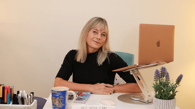 A woman sits at a desk with a laptop, pens, and a plant.