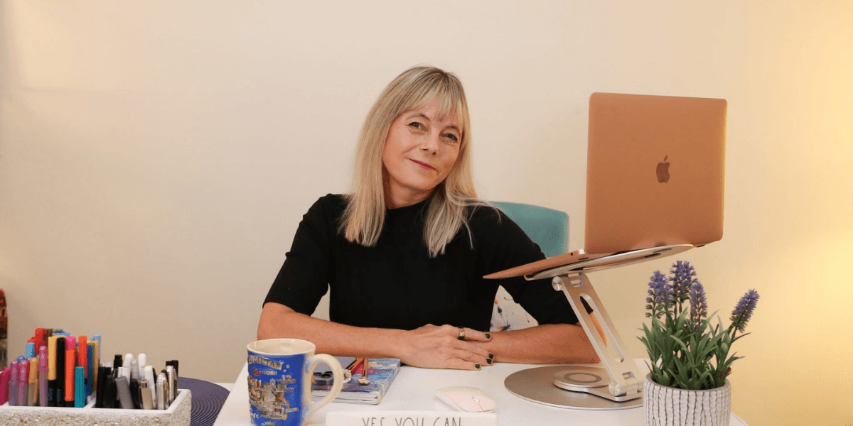 A woman sits at a desk with a laptop, pens, and a plant.