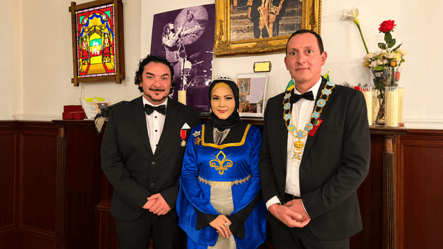 Three individuals in formal attire posed indoors with decorations and a historical backdrop