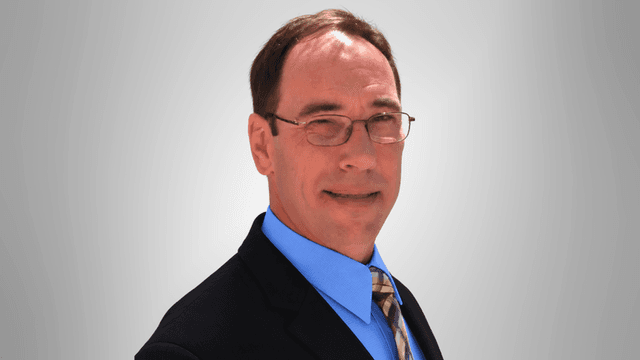 Man in a suit with glasses, smiling against a gray background