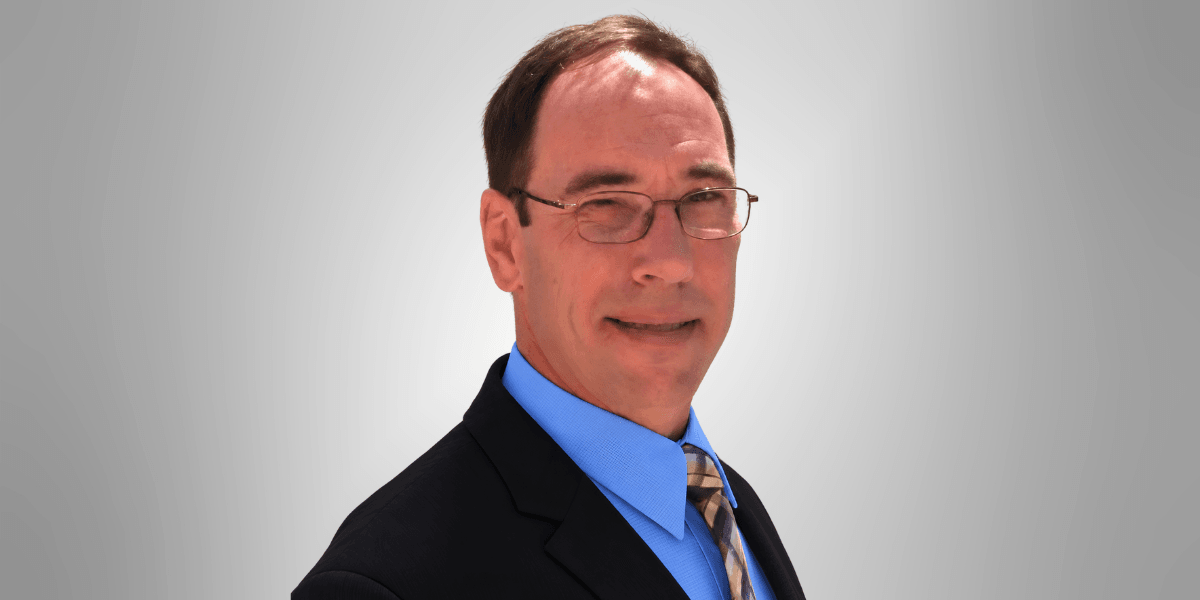 Man in a suit with glasses, smiling against a gray background