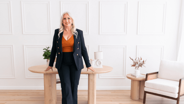 A blonde woman in a stylish outfit stands confidently at a wooden table.