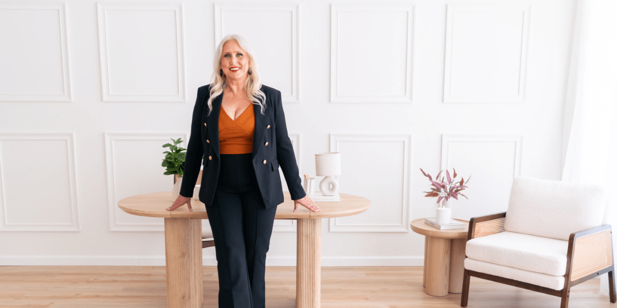 A blonde woman in a stylish outfit stands confidently at a wooden table.