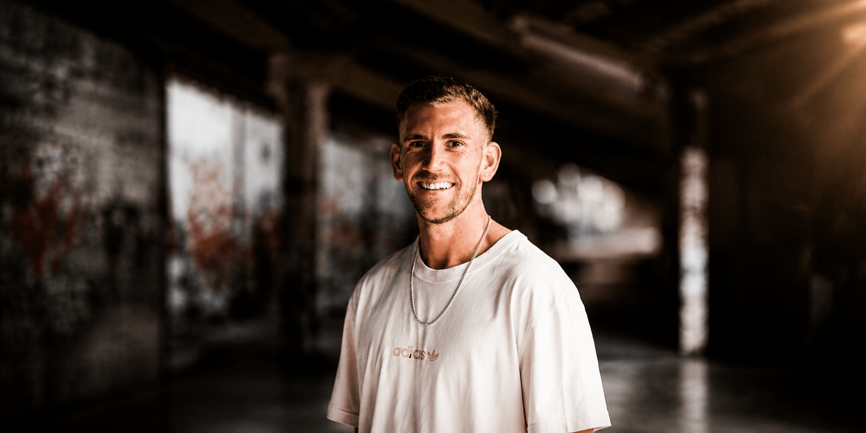 Smiling man in a casual white shirt stands in a graffiti-covered urban setting