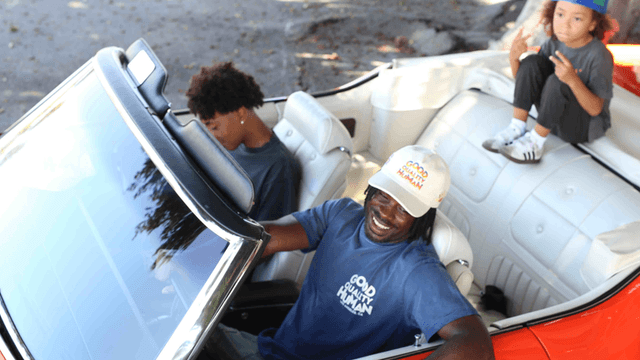 A smiling man drives a classic convertible with two kids inside.