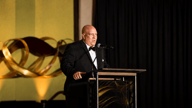 A man in a tuxedo speaks at a podium during an event.