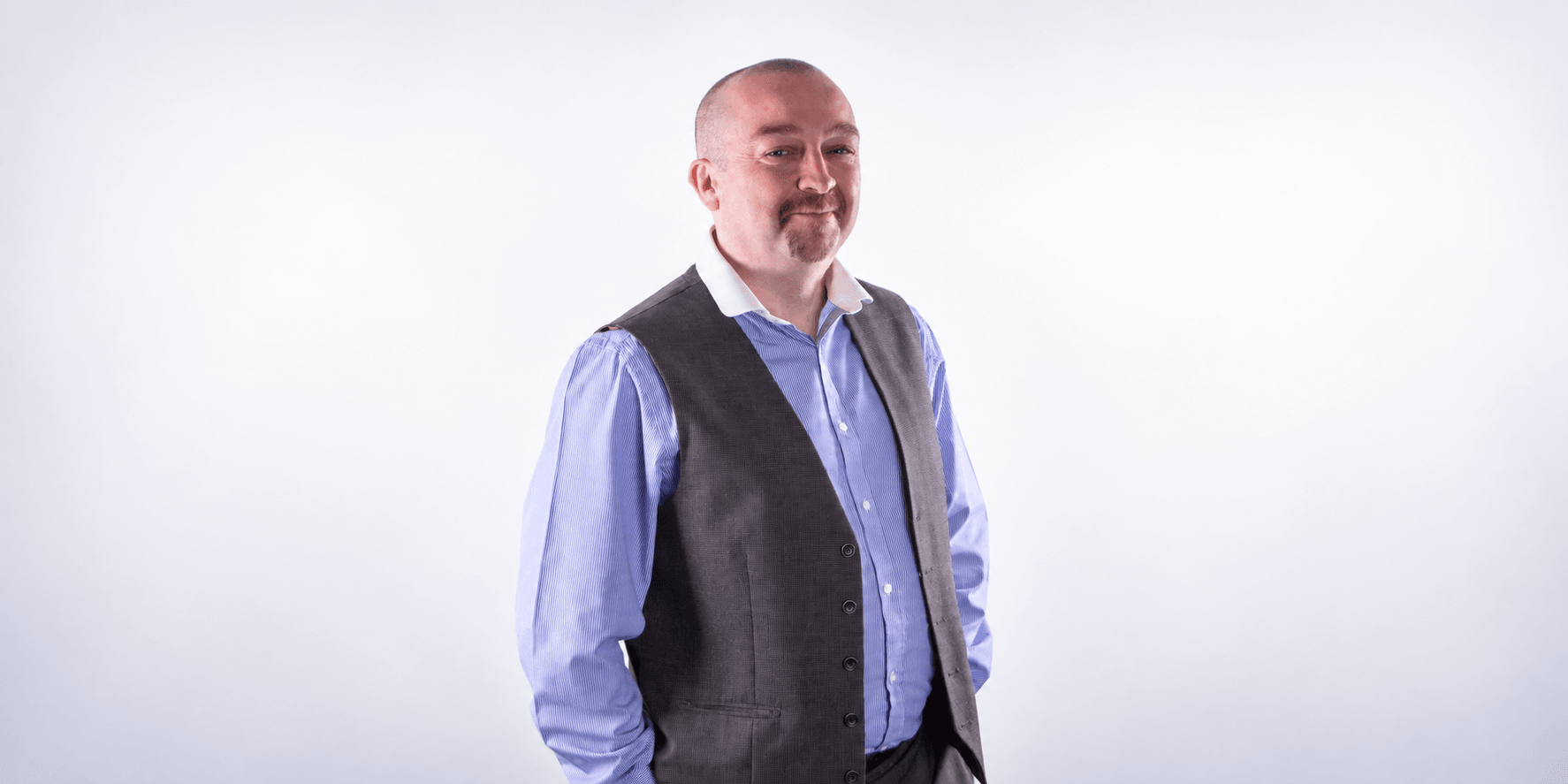 Man in a grey vest and blue shirt standing against a neutral background
