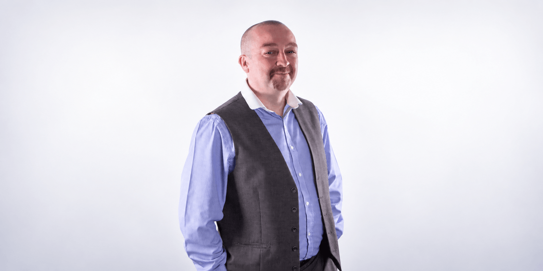 Man in a grey vest and blue shirt standing against a neutral background