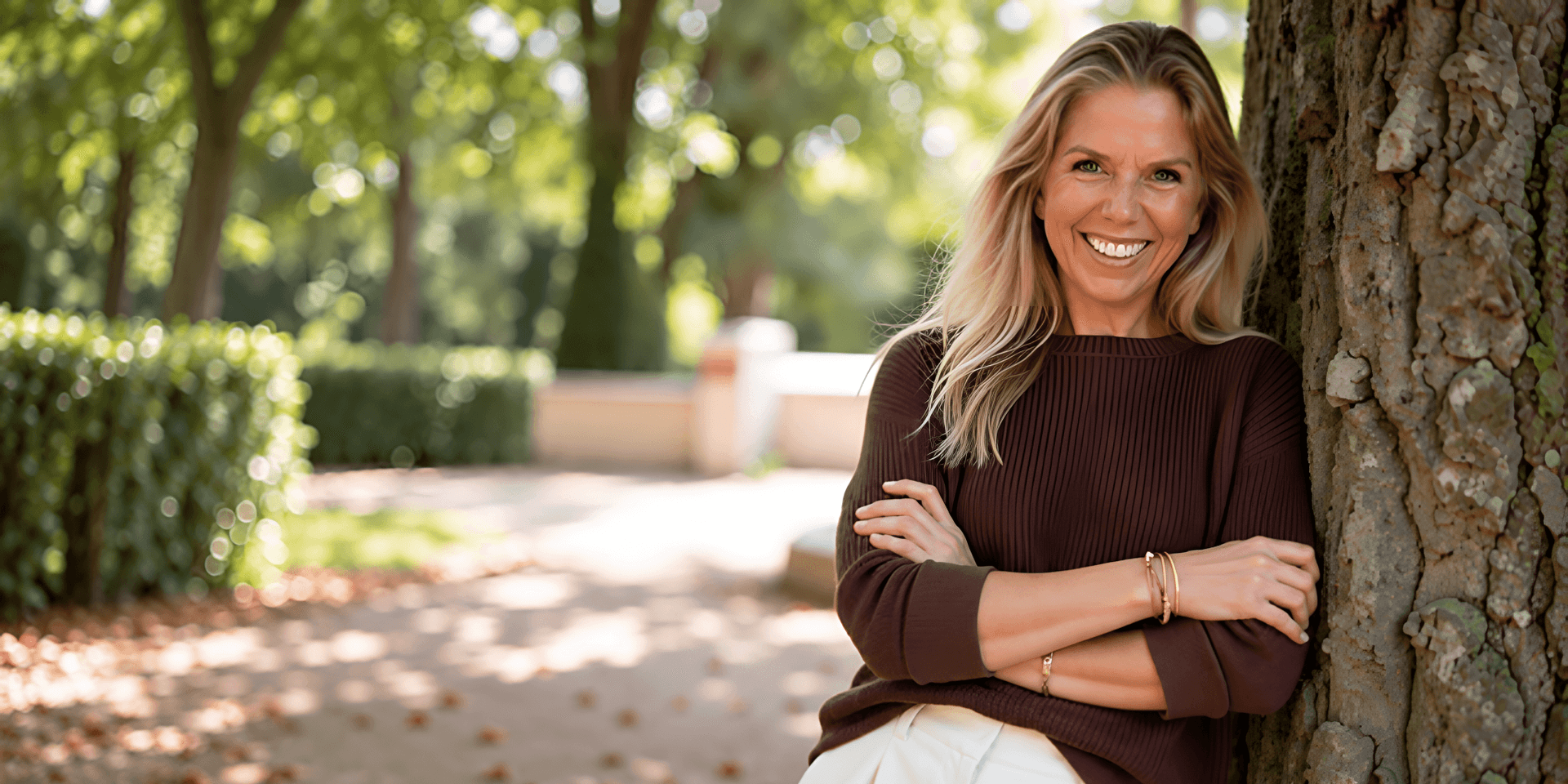 Smiling woman in a brown sweater standing against a tree in a park