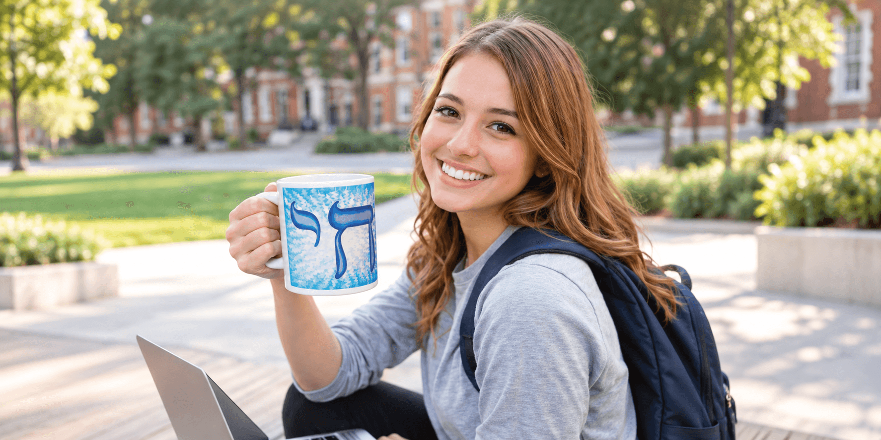 Young woman sitting outdoors, holding a coffee mug, smiling, with a laptop beside her