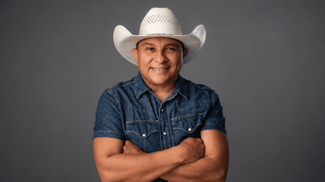 Man in a cowboy hat and denim shirt poses with arms crossed against a gray backdrop