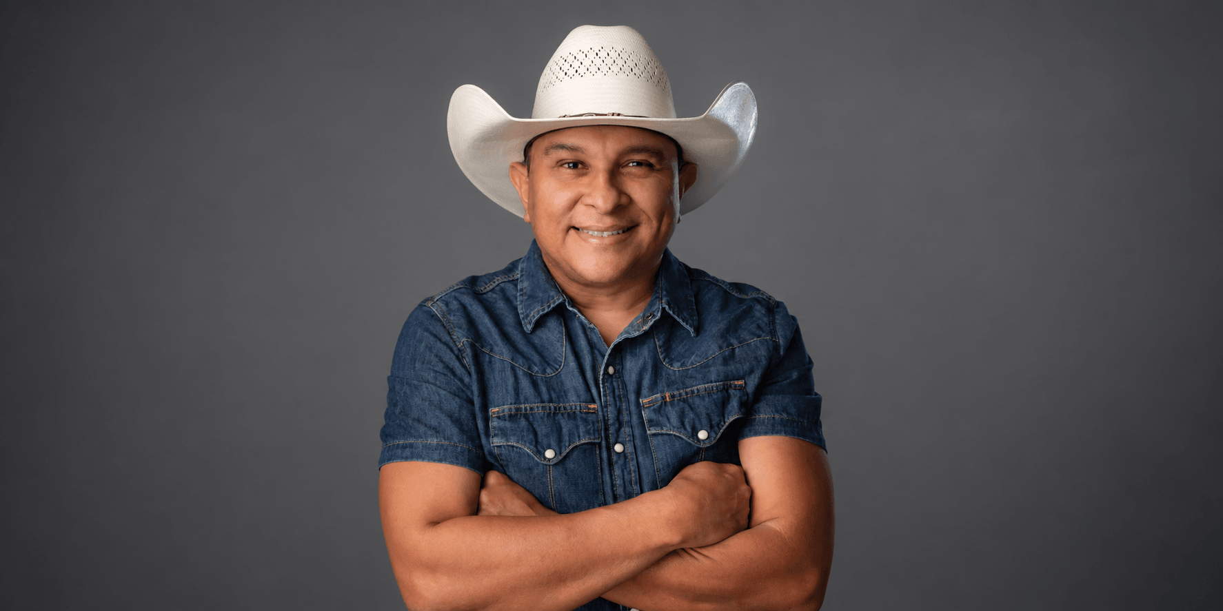 Man in a cowboy hat and denim shirt poses with arms crossed against a gray backdrop