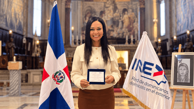 A woman holds a certificate, standing between the Dominican Republic and INEX flags indoors