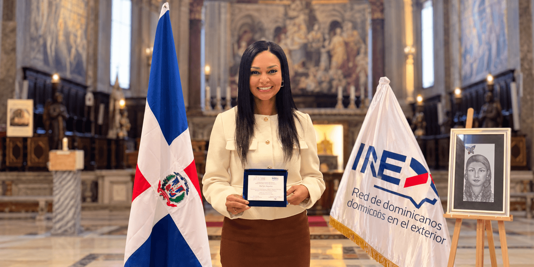 A woman holds a certificate, standing between the Dominican Republic and INEX flags indoors