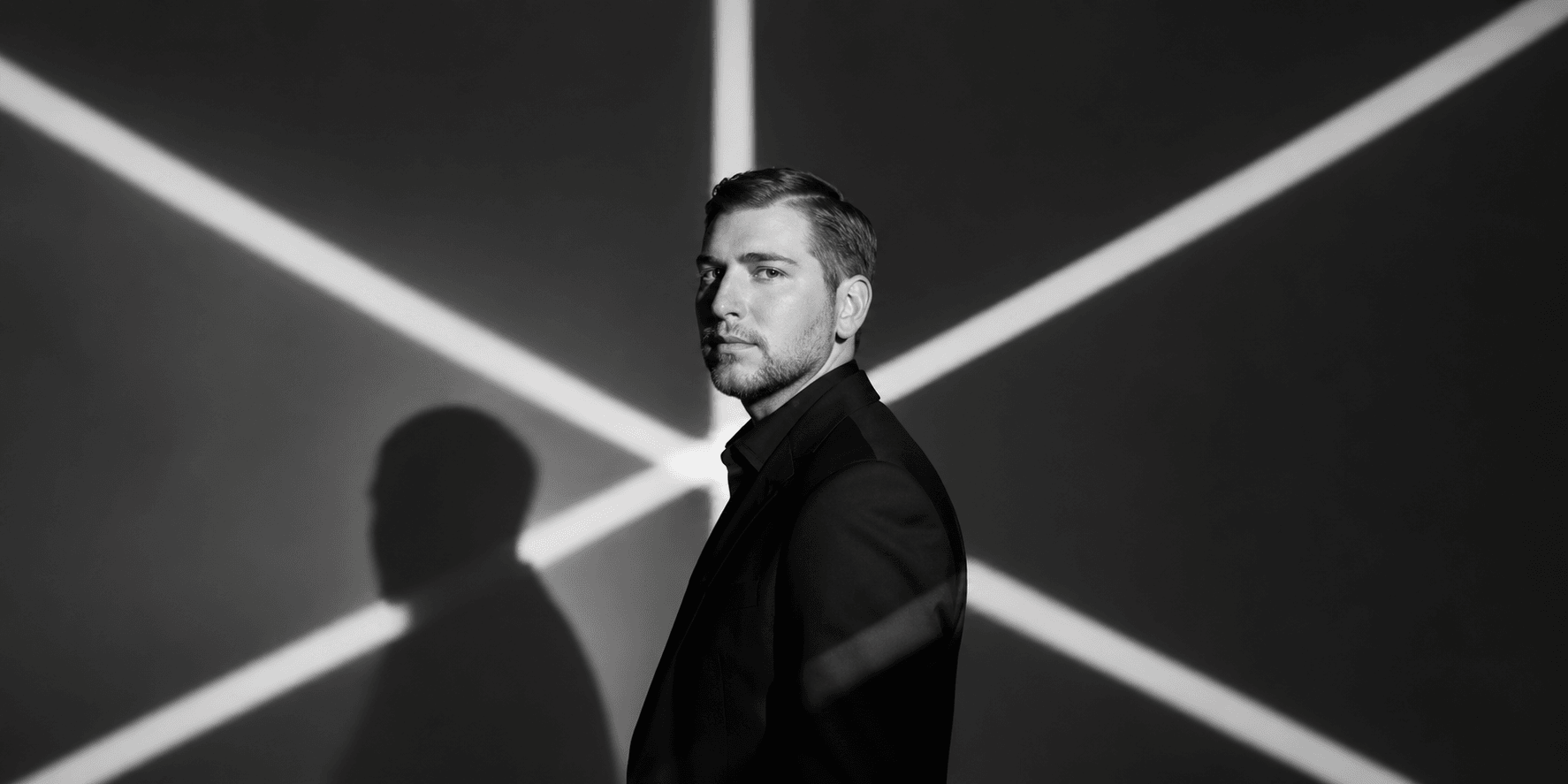 A man in a suit poses against a stark black and white background with shadows
