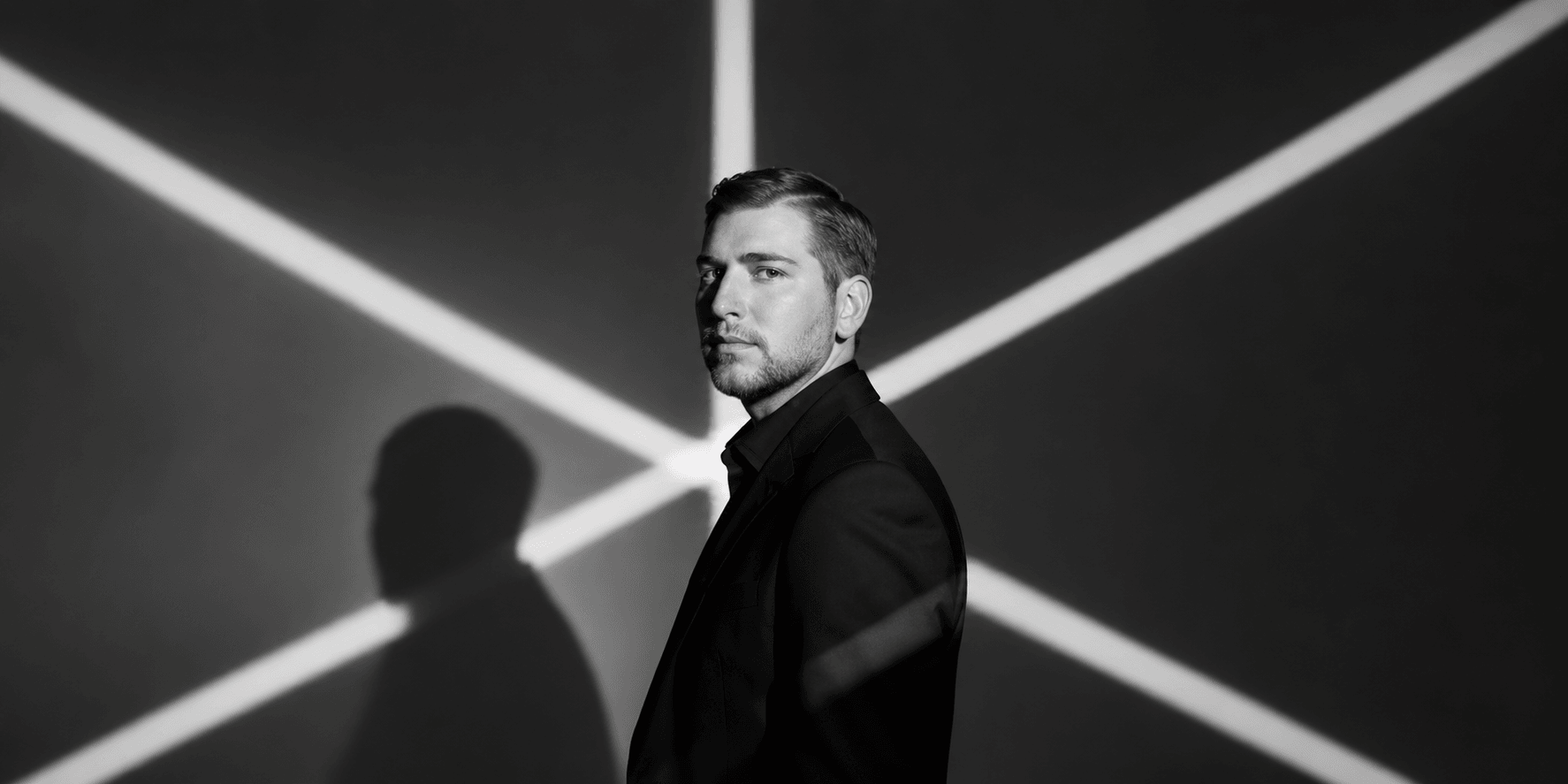 A man in a suit poses against a stark black and white background with shadows