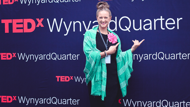 Person in a green shawl posing with a flower at a TEDx event backdrop